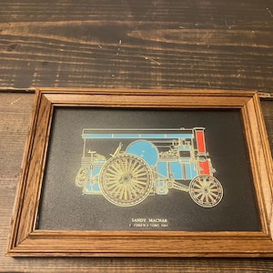 May include: Framed artwork featuring a detailed illustration of a steam engine in blue, red, and gold against a black background. The artwork is in a wooden frame. The text "SANDY MACNAB" is visible.
