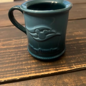May include: A teal ceramic mug with a handle. The mug features a raised design of the Disney Cruise Line logo and the words "Disney Cruise Line" around the base. The mug is sitting on a dark wooden surface.