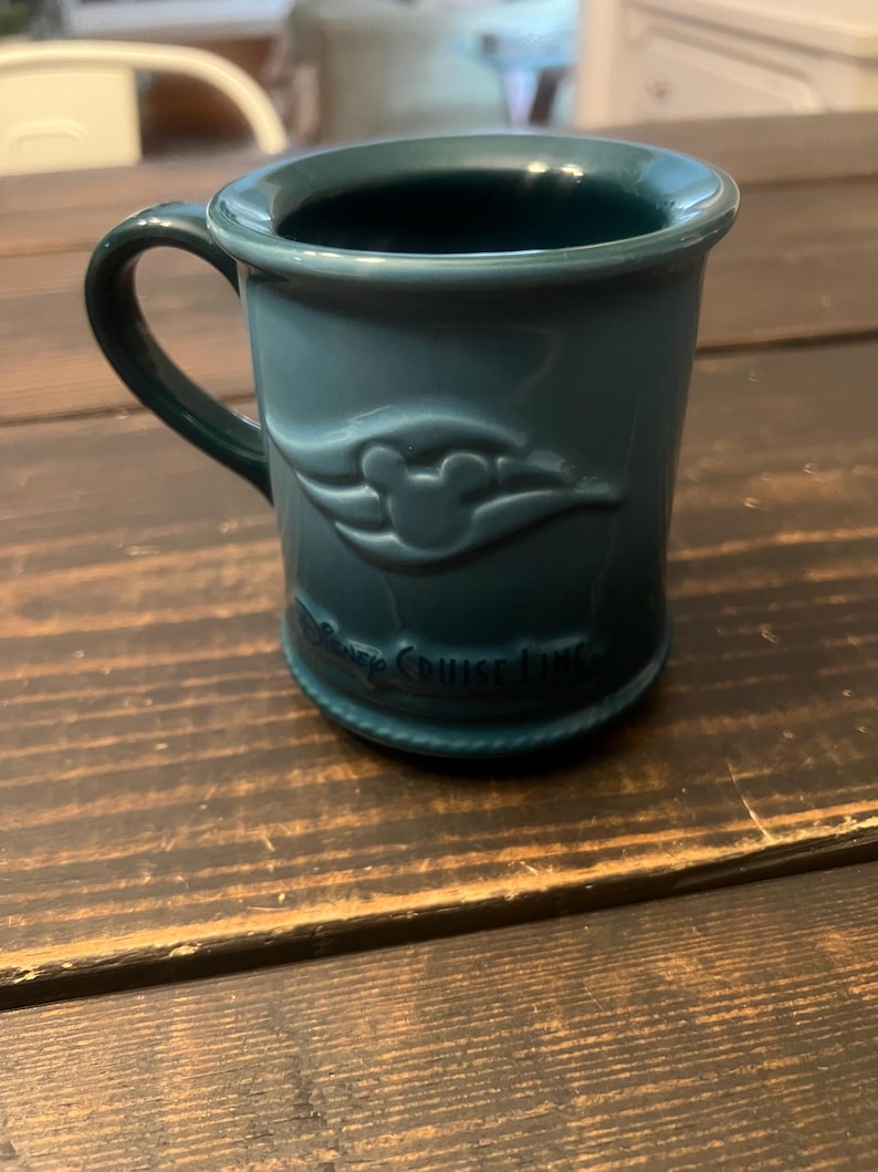 May include: A teal ceramic mug with a handle. The mug features a raised design of the Disney Cruise Line logo and the words "Disney Cruise Line" around the base. The mug is sitting on a dark wooden surface.