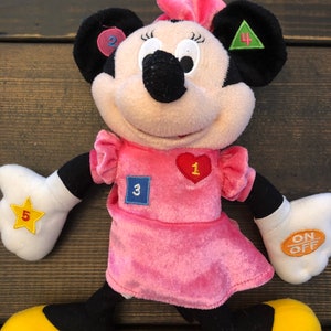 May include: A pink plush Minnie Mouse toy with a pink dress and yellow shoes. She has a heart with the number 1, a blue square with the number 3, a green triangle with the number 4, a yellow star with the number 5, and a pink circle with the number 2 on her. She also has an on/off switch on her arm.