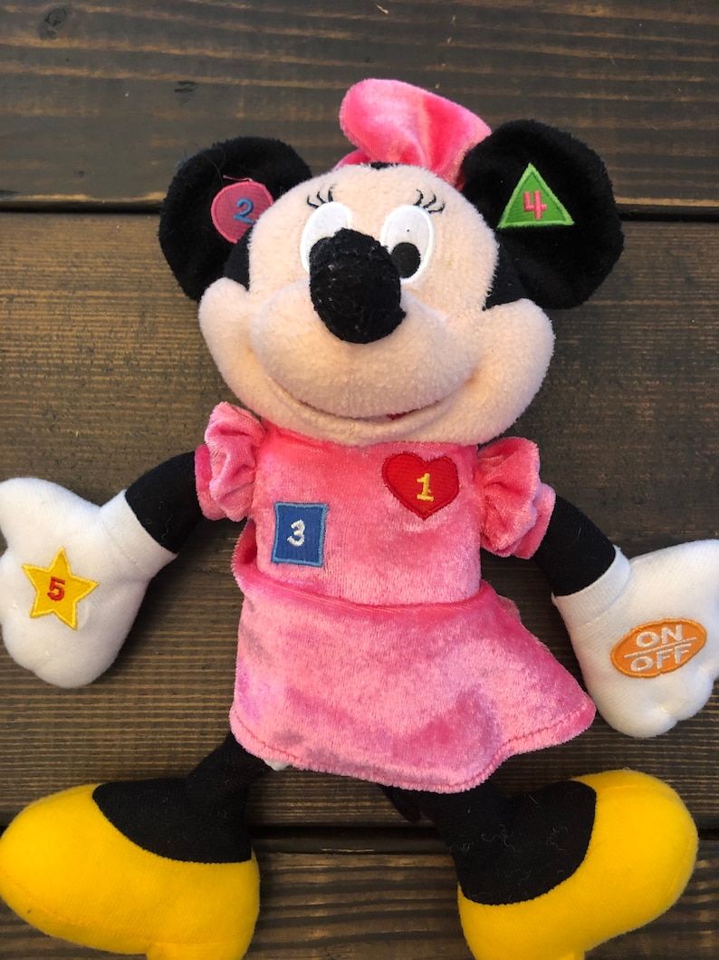 May include: A pink plush Minnie Mouse toy with a pink dress and yellow shoes. She has a heart with the number 1, a blue square with the number 3, a green triangle with the number 4, a yellow star with the number 5, and a pink circle with the number 2 on her. She also has an on/off switch on her arm.