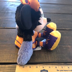 May include: A plush Minnie Mouse doll in a Halloween costume. The doll has a black body, a white face, and a bow with orange and purple accents. The doll is sitting on a wooden surface.