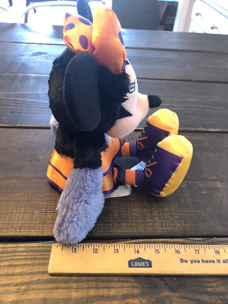May include: A plush Minnie Mouse doll in a Halloween costume. The doll has a black body, a white face, and a bow with orange and purple accents. The doll is sitting on a wooden surface.