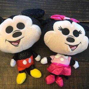May include: A pair of plush Mickey Mouse and Minnie Mouse toys. Mickey is wearing red shorts and yellow shoes. Minnie is wearing a pink dress and red shoes. Both toys have black sparkly ears and a black sparkly body.
