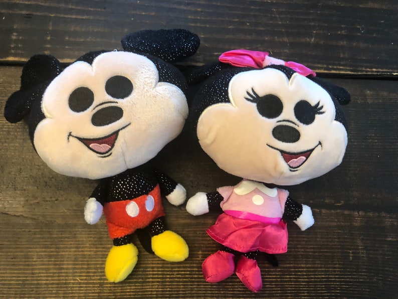 May include: A pair of plush Mickey Mouse and Minnie Mouse toys. Mickey is wearing red shorts and yellow shoes. Minnie is wearing a pink dress and red shoes. Both toys have black sparkly ears and a black sparkly body.