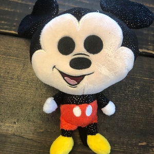 May include: A plush Mickey Mouse toy with black ears, a black nose, and red shorts with white buttons. The toy has a sparkly black and white design.