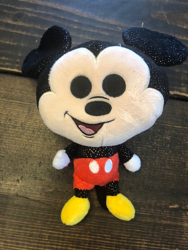 May include: A plush Mickey Mouse toy with black ears, a black nose, and red shorts with white buttons. The toy has a sparkly black and white design.