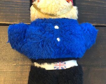 May include: A plush toy bear dressed as a British soldier. The bear is wearing a black hat with a red and black plaid band, a blue jacket with two white buttons, black pants, and a white belt with a Union Jack patch. The bear has a gold chain around its neck.