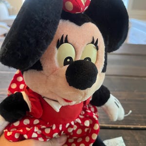 May include: A plush Minnie Mouse toy wearing a red dress with white polka dots and a red and white polka dot bow. The toy has black eyes with white eyelashes and a black nose.