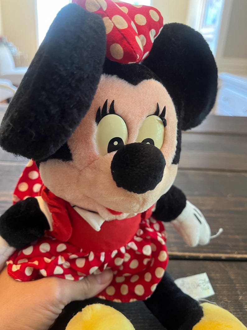May include: A plush Minnie Mouse toy wearing a red dress with white polka dots and a red and white polka dot bow. The toy has black eyes with white eyelashes and a black nose.