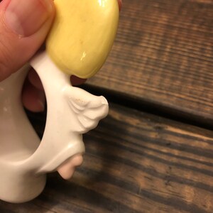 Puede incluir: Figurilla de &aacute;ngel de cer&aacute;mica blanca con cabello amarillo y un ala. El &aacute;ngel sostiene un anillo en su mano.