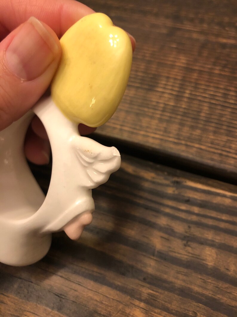 Puede incluir: Figurilla de &aacute;ngel de cer&aacute;mica blanca con cabello amarillo y un ala. El &aacute;ngel sostiene un anillo en su mano.