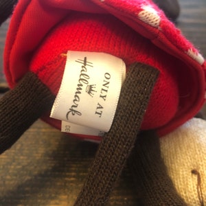 K&ouml;nnte beinhalten: Rotes und wei&szlig;es gestricktes Pl&uuml;schtier mit braunen gestrickten Armen und einem wei&szlig;en Etikett mit der Aufschrift "Hallmark Only At" mit einem Kronenlogo.