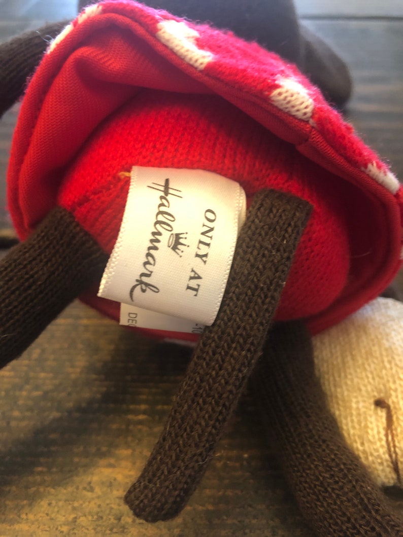 K&ouml;nnte beinhalten: Rotes und wei&szlig;es gestricktes Pl&uuml;schtier mit braunen gestrickten Armen und einem wei&szlig;en Etikett mit der Aufschrift "Hallmark Only At" mit einem Kronenlogo.