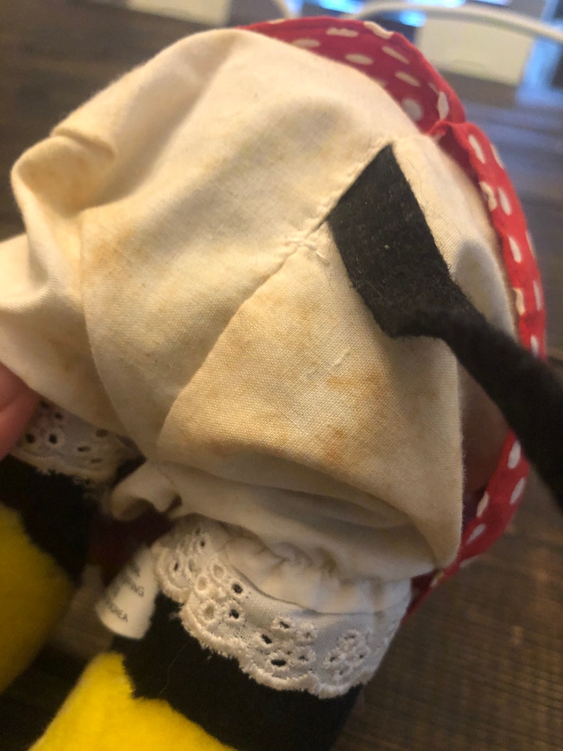 May include: A plush bee toy with yellow and black stripes, wearing a white dress with red polka dots and a black bow. The dress has a white lace trim and the bee has a black and white striped tail.