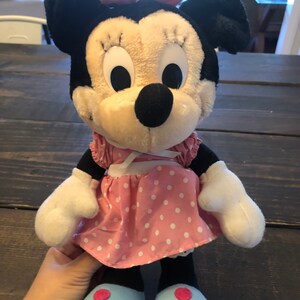 May include: A plush toy of Minnie Mouse wearing a pink polka dot dress with a white bow. She has black ears, a black nose, and large white eyes.