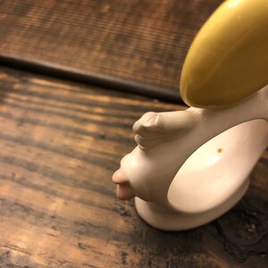 Puede incluir: Anillo de servilleta de cer&aacute;mica blanca con una cara amarilla y rosa. El anillo tiene forma de cabeza humana con un halo amarillo.
