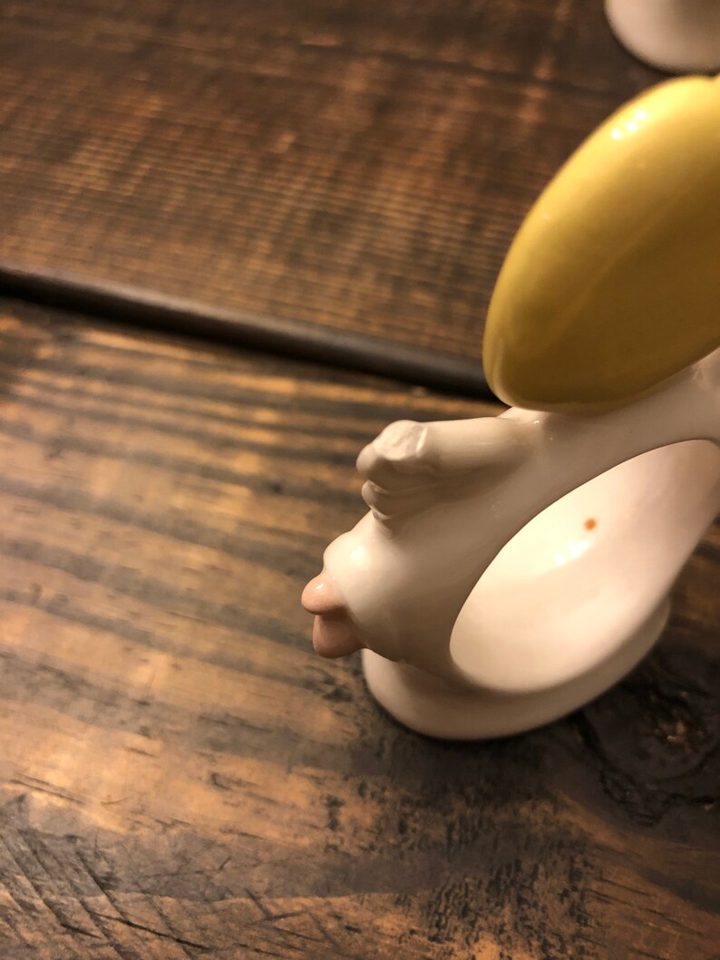 Puede incluir: Anillo de servilleta de cer&aacute;mica blanca con una cara amarilla y rosa. El anillo tiene forma de cabeza humana con un halo amarillo.