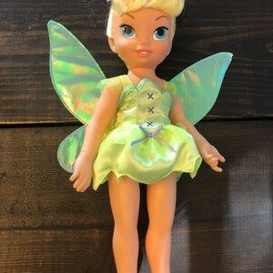 May include: A green and iridescent fairy doll with blonde hair and a green dress. The doll has a bow on its head and a pair of iridescent wings.