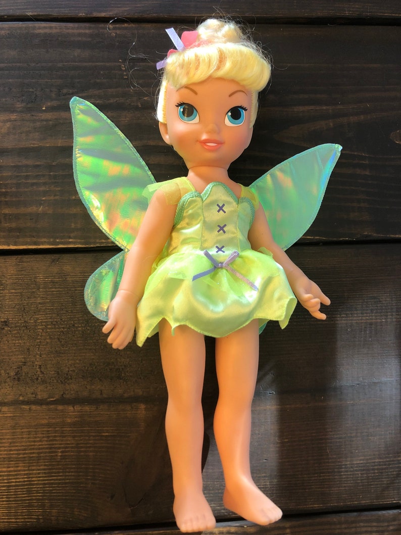 May include: A green and iridescent fairy doll with blonde hair and a green dress. The doll has a bow on its head and a pair of iridescent wings.