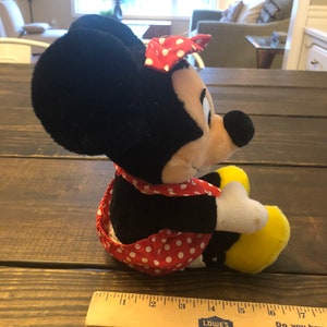 May include: A plush toy of Minnie Mouse wearing a red and white polka dot dress and a red bow. The toy is sitting on a wooden surface with a ruler next to it. The ruler is marked with inches and has the words "Lowe's" and "Do you have a...