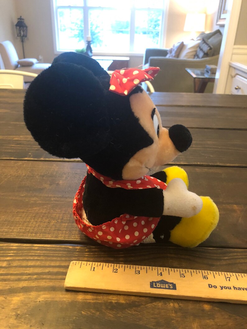 May include: A plush toy of Minnie Mouse wearing a red and white polka dot dress and a red bow. The toy is sitting on a wooden surface with a ruler next to it. The ruler is marked with inches and has the words "Lowe's" and "Do you have a...
