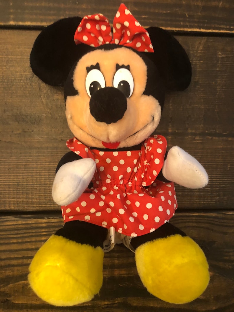 May include: A plush toy of Minnie Mouse wearing a red dress with white polka dots and a red bow. She has black ears, a black nose, and yellow shoes.