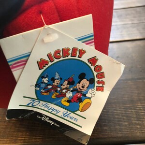 Peut inclure: Une &eacute;tiquette blanche avec un cercle bleu repr&eacute;sentant Mickey Mouse et ses amis. L'&eacute;tiquette indique "Mickey Mouse 70 Happy Years" et "The Disney Store".