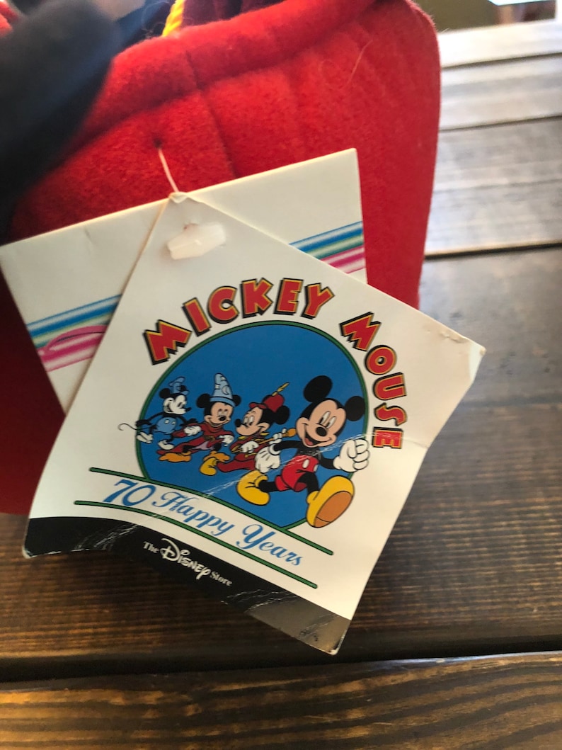 Peut inclure: Une &eacute;tiquette blanche avec un cercle bleu repr&eacute;sentant Mickey Mouse et ses amis. L'&eacute;tiquette indique "Mickey Mouse 70 Happy Years" et "The Disney Store".