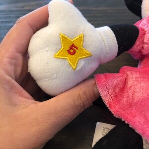 May include: A pink and black plush toy with a white glove. The glove has a yellow star with a red number 5 embroidered on it.