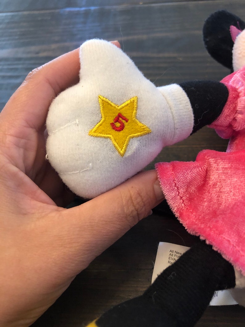 May include: A pink and black plush toy with a white glove. The glove has a yellow star with a red number 5 embroidered on it.