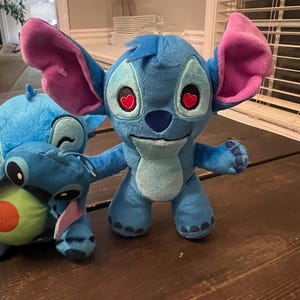 Op de afbeelding: Een blauwe Stitch pluche pop met grote roze gevoerde oren en hartvormige ogen. Een andere Stitch pluche pop is gedeeltelijk zichtbaar aan de linkerkant, samen met een avocado pluche. De poppen staan op een houten oppervlak.