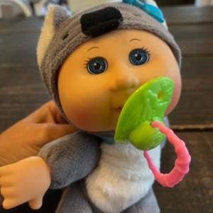 May include: A gray plush koala baby doll with blue eyes and a green and pink pacifier. The doll is wearing a gray koala costume with a blue bow.