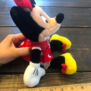 May include: A red and black plush Minnie Mouse toy with yellow shoes and a red polka dot dress. The toy is sitting on a wooden surface with a ruler next to it. The ruler is marked with inches and has the word "LOWE'S" printed on it.