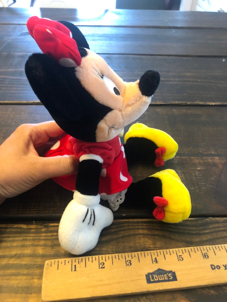 May include: A red and black plush Minnie Mouse toy with yellow shoes and a red polka dot dress. The toy is sitting on a wooden surface with a ruler next to it. The ruler is marked with inches and has the word "LOWE'S" printed on it.