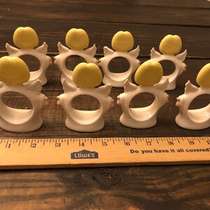 Puede incluir: Conjunto de ocho anillos de servilleta de cer&aacute;mica blanca con alas de &aacute;ngel amarillas y un c&iacute;rculo amarillo en la parte superior. Los anillos est&aacute;n dispuestos en una fila sobre una superficie de madera.