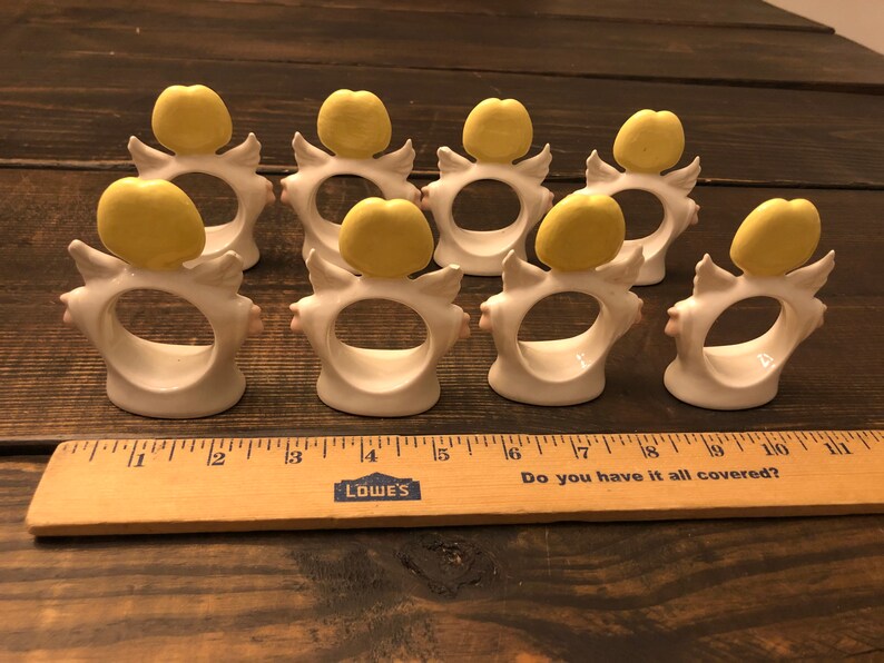 Puede incluir: Conjunto de ocho anillos de servilleta de cer&aacute;mica blanca con alas de &aacute;ngel amarillas y un c&iacute;rculo amarillo en la parte superior. Los anillos est&aacute;n dispuestos en una fila sobre una superficie de madera.