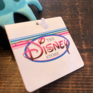 May include: A white tag with the text "The Disney Store" in a pink, blue, and white circle. The tag has a rainbow striped background.