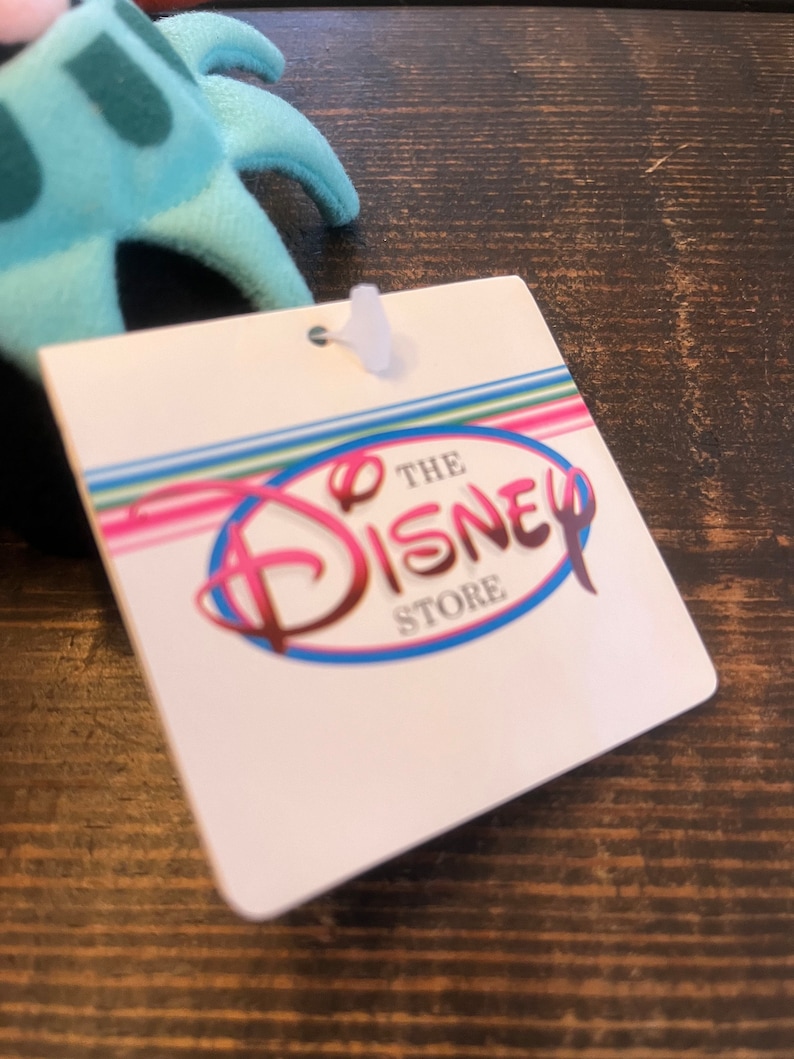 May include: A white tag with the text "The Disney Store" in a pink, blue, and white circle. The tag has a rainbow striped background.