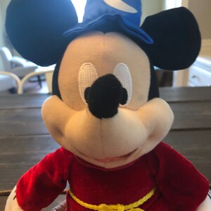 May include: A plush Mickey Mouse wearing a red robe with a yellow rope belt and a blue wizard hat with a white crescent moon.