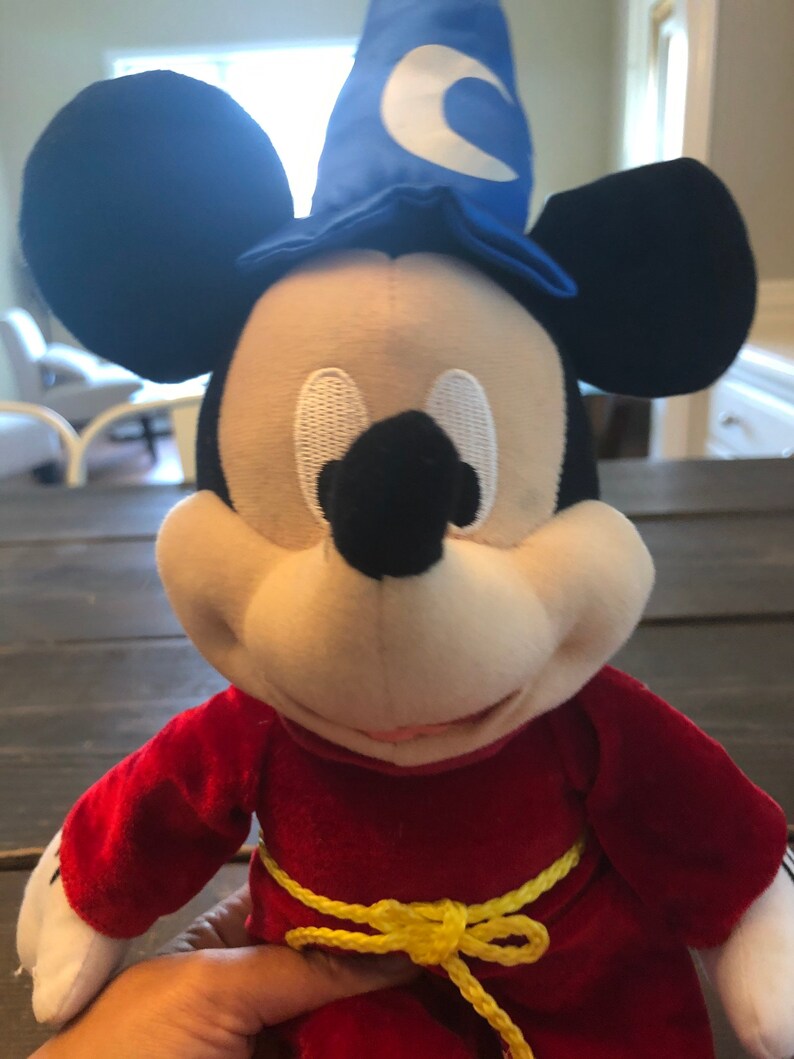 May include: A plush Mickey Mouse wearing a red robe with a yellow rope belt and a blue wizard hat with a white crescent moon.