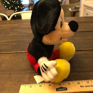May include: A black and red plush Mickey Mouse toy sitting on a wooden surface. The toy is about 15 inches tall and has a yellow nose and white gloves.