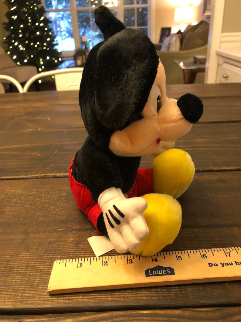 May include: A black and red plush Mickey Mouse toy sitting on a wooden surface. The toy is about 15 inches tall and has a yellow nose and white gloves.