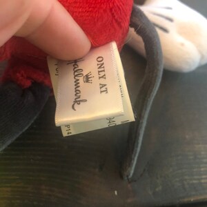 Puede incluir: Un juguete de peluche de Mickey Mouse con cabeza roja, cuerpo negro y guantes blancos. La etiqueta del juguete dice "Only at Hallmark" con un logotipo de corona.