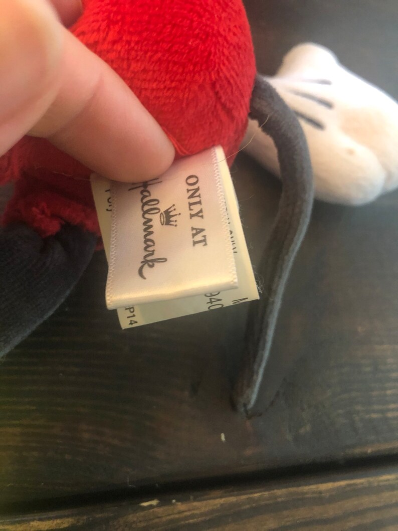 Puede incluir: Un juguete de peluche de Mickey Mouse con cabeza roja, cuerpo negro y guantes blancos. La etiqueta del juguete dice "Only at Hallmark" con un logotipo de corona.