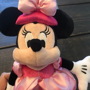 May include: A pink and black plush toy of Minnie Mouse wearing a pink dress with white polka dots and a pink hat with a pink bow.
