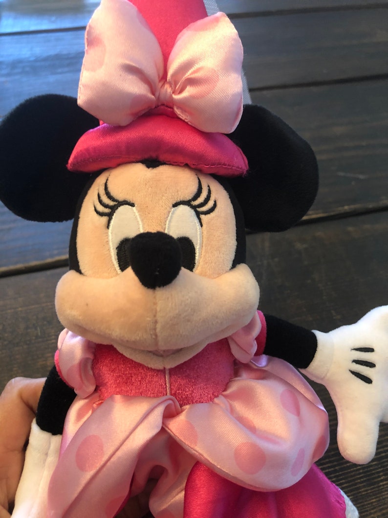 May include: A pink and black plush toy of Minnie Mouse wearing a pink dress with white polka dots and a pink hat with a pink bow.