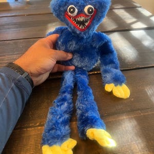 May include: A blue plush toy with a triangular head, large eyes, and a wide red mouth filled with white teeth. The toy has long arms and legs with yellow hands and feet. The toy is made of soft, fuzzy material.
