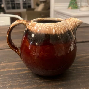 May include: A brown and white ceramic creamer with a brown glaze. The creamer has a rounded body and a handle.