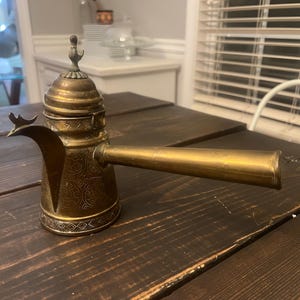 May include: An antique brass coffee pot with an ornate design. The pot features a long spout and a decorative lid with a small handle. The pot is sitting on a dark wooden table.